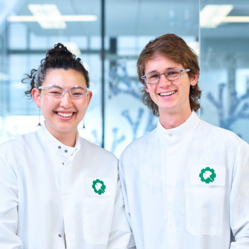 A photo of two people both wearing white lab coats with a green Ginkgo Bioworks logo on the chest and both wearing safety goggles, standing in front of a glass wall of a lab