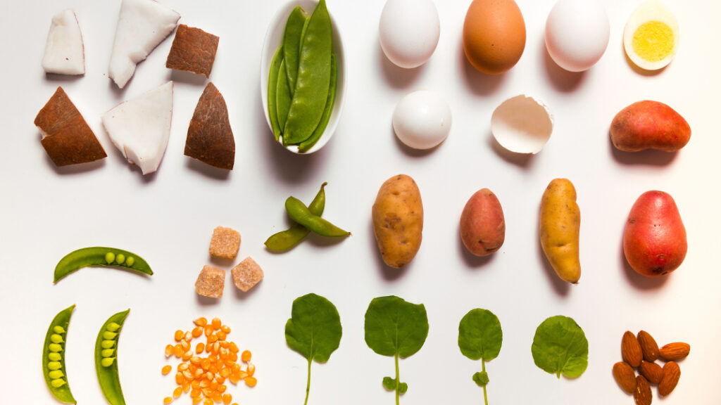 An overhead shot of an organized array of foods laid out on a white surface, the foods include eggs, potatoes, coconut, legumes, nuts, sugar, and seeds