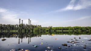 A polluted waterway showing garbage in the foreground floating in shallow water with large distillation stacks in the distance implying that the water is contaminated