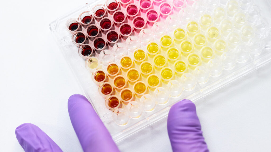 A scientist's gloved hand holds a 96-well plate with a colorful array of samples